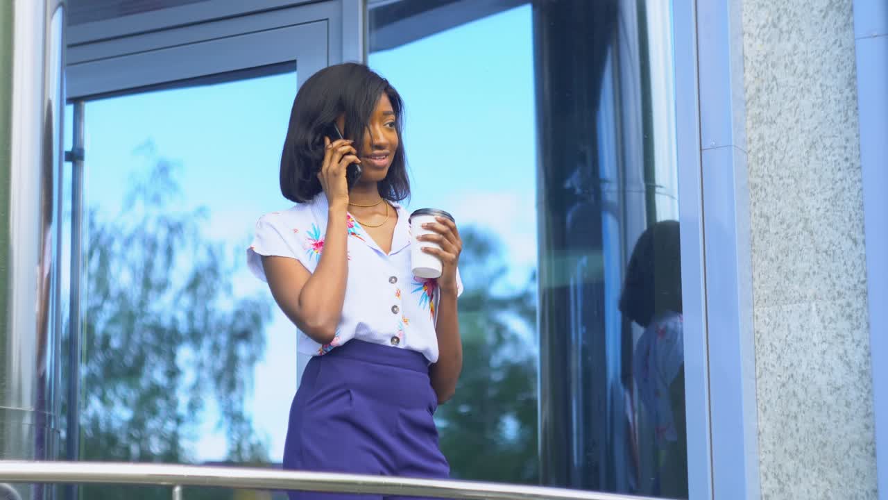 una joven afroamericana adulta está hablando por teléfono afuera cerca de un edificio de oficinas moderno.