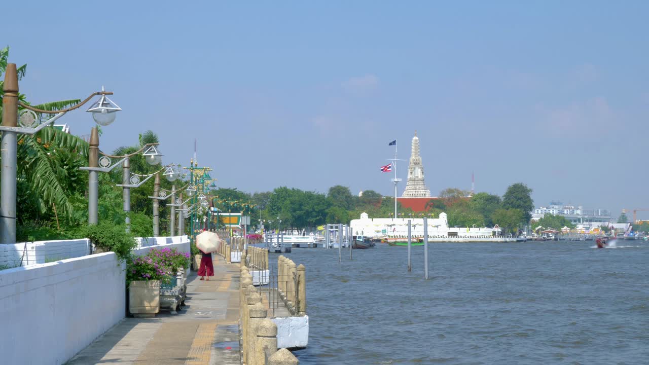 Una donna con un ombrello bianco cammina lungo la passeggiata del lungofiume vicino a Wat Arun, Thailandia, con un motoscafo tradizionale che attraversa il fiume - Inquadratura ampia