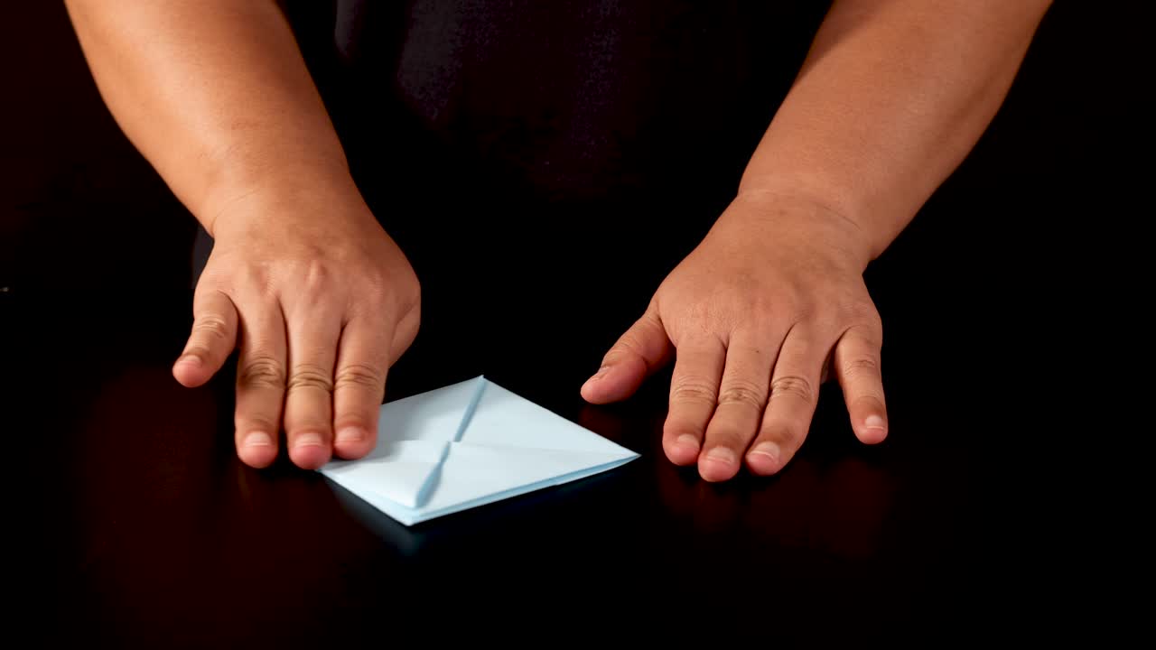 Person folds blue paper into origami boat, overhead view, dark background, even studio lighting