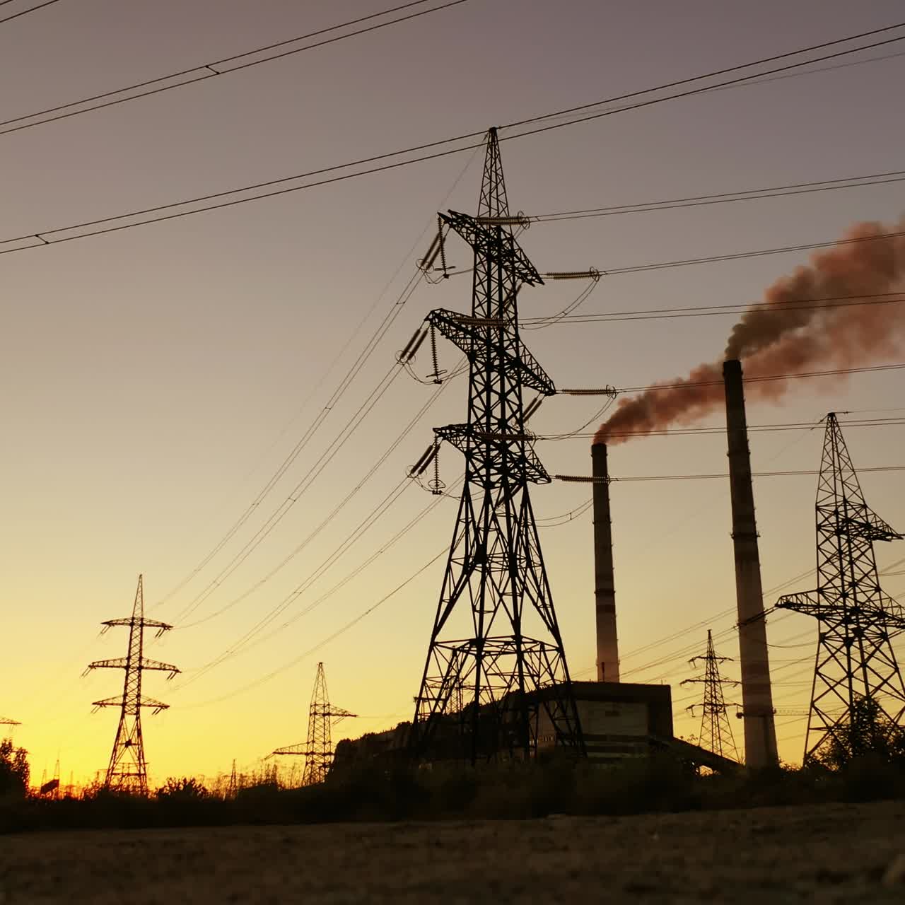 High voltage electric lines and industry. Pipes with dark smoke near the transmission electric towers at dusk. Pollution the atmosphere. View from below.