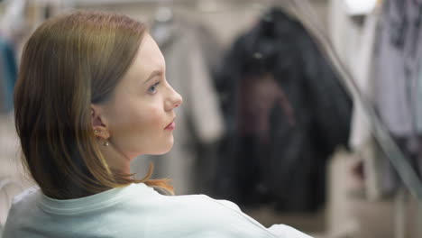 close-up of young woman with thoughtful expression seated indoors in a mall, showcasing soft lighting, subtle focus on hairstyle, and blurred background featuring modern clothing displays