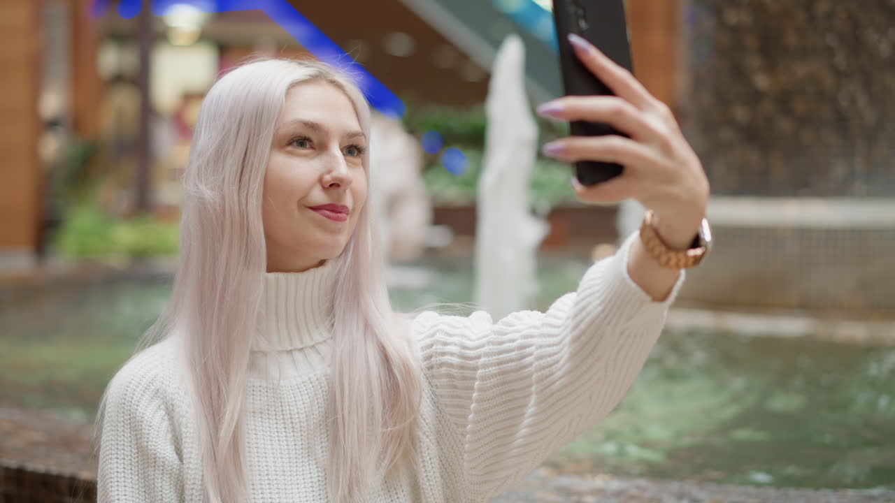 Cheerful mall visitor sits beside water fountain on bench adjusting hair and posing with mobile phone, capturing sparkling water jets under bright mall lights while shoppers pass by
