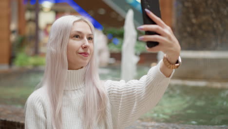 cheerful mall visitor sits beside water fountain on bench adjusting hair and posing with mobile phone, capturing sparkling water jets under bright mall lights while shoppers pass by