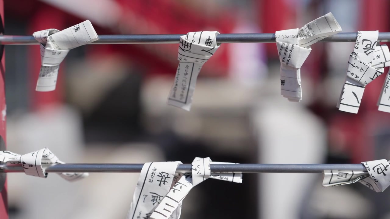 Rullede omikuji-spådomspapirer med kanji bundet til metalstænger ved en helligdom med sløret menneskemængde i baggrunden, Japan.