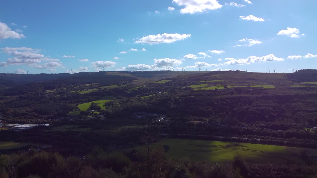 Aerial Rising Over Welsh Valley with Open Fields and Roads Below with Wind Turbine Farm in Background on Hilltop. Clear Blue Sky