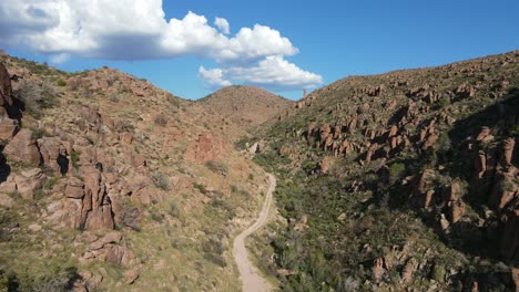 Cinematic-aerial-shot,-drone-flying-over-dirt-road-at-the-bottom-of-a-jagged-canyon-on-a-bright-sunny-day,-blue-skies