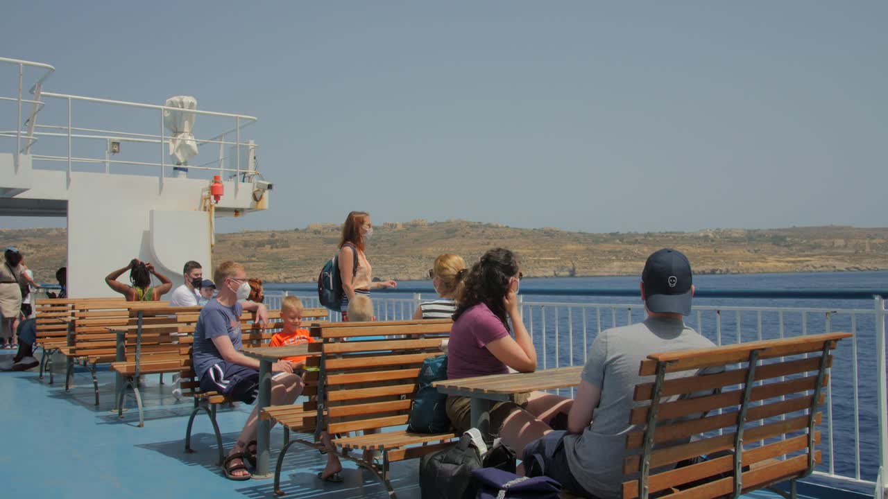 Tourists In Masks Viewing Seascape From Channel Ferry Cruising Across The Sea In Gozo, Malta During Covid Pandemic. wide shot