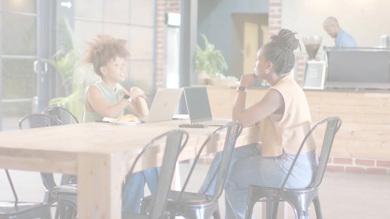 Two women leaning at table tapping wrist, causing sporadic glitch lines over laptops during work