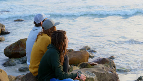 Side-view-of-young-caucasian-friends-sitting-on-rock-and-looking-at-sea-on-the-beach-4k