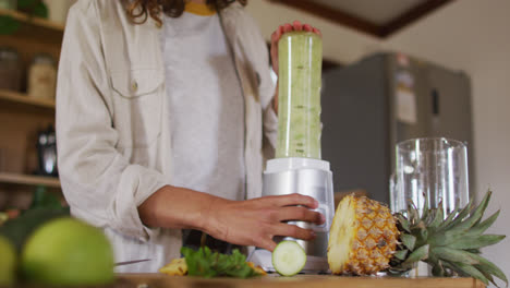 Happy-mixed-race-woman-blending-health-drink-standing-in-cottage-kitchen-smiling