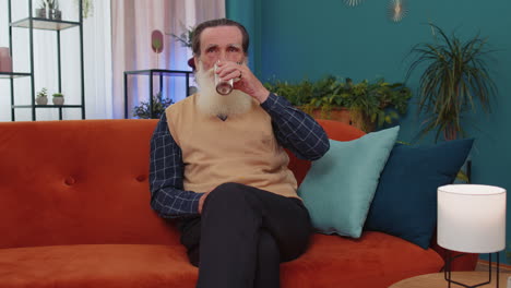anciano anciano sediento sentado en casa sosteniendo un vaso de agua natural hacer sorbos bebiendo agua tranquila