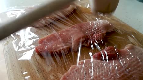 ablandamiento del lomo de cerdo con un martillo de carne de madera a través de una película adhesiva sobre una tabla de cortar de madera, vista lateral slomo