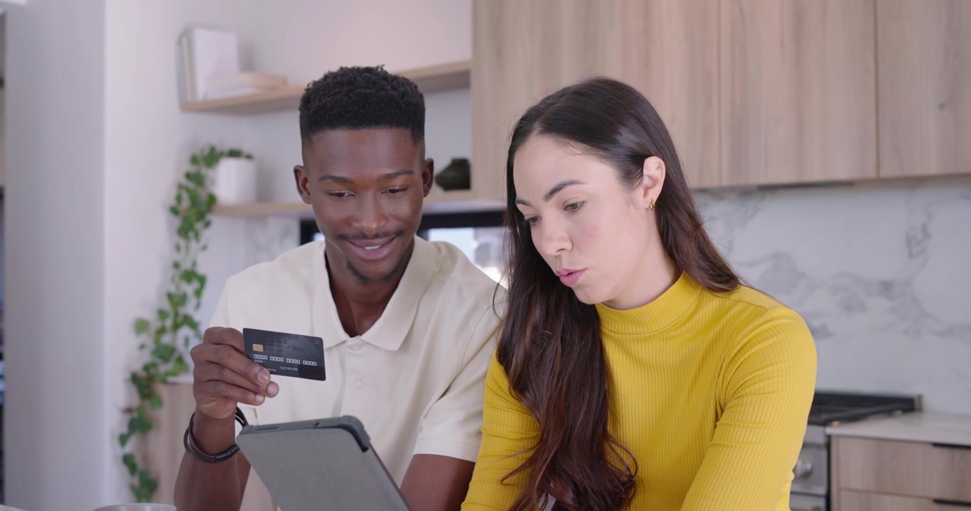 Diverse couple sitting at marble counter, woman in yellow tapping tablet, man pulling credit card