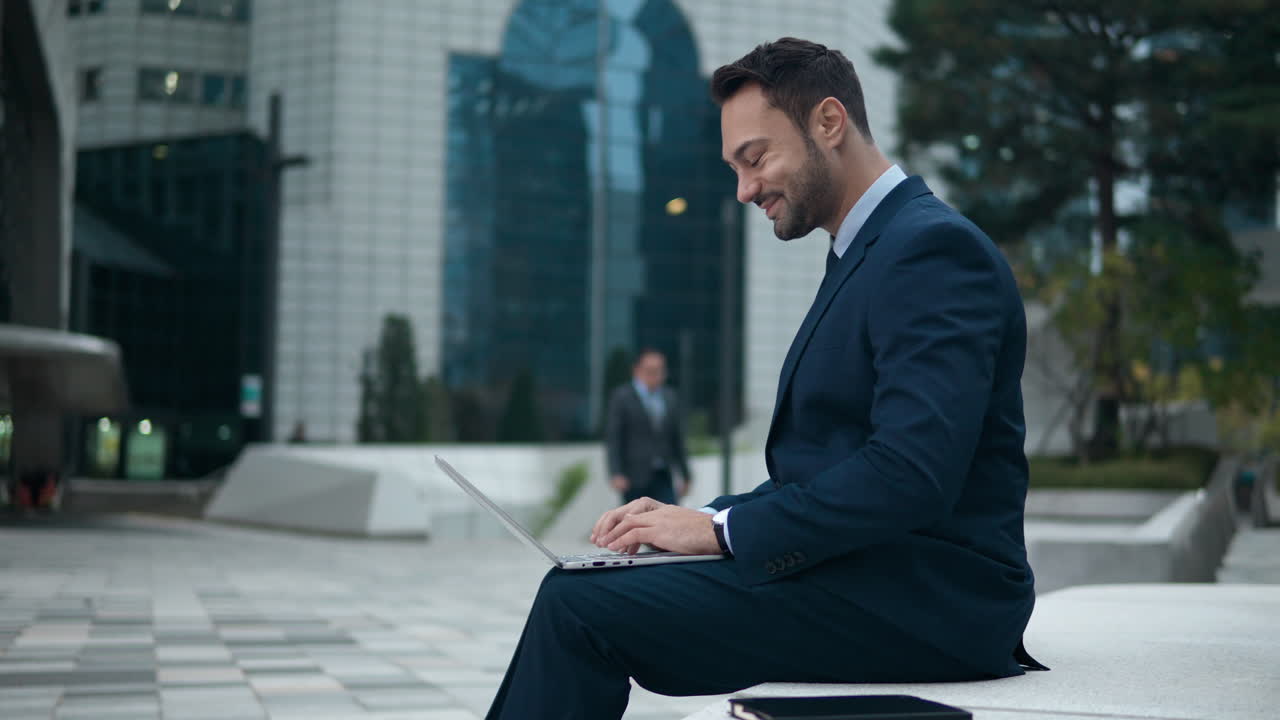 hombre de negocios feliz trabajando escribiendo en una computadora portátil fuera del edificio de oficinas sentado en un banco
