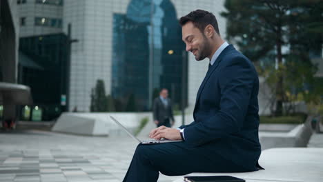happy businessman working typing on laptop outside office building sitting on bench