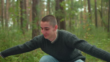 young student in dark green knitted sweater smiling warmly while preparing tent in woodland setting, surrounded by tall trees and green foliage during peaceful outdoor camping