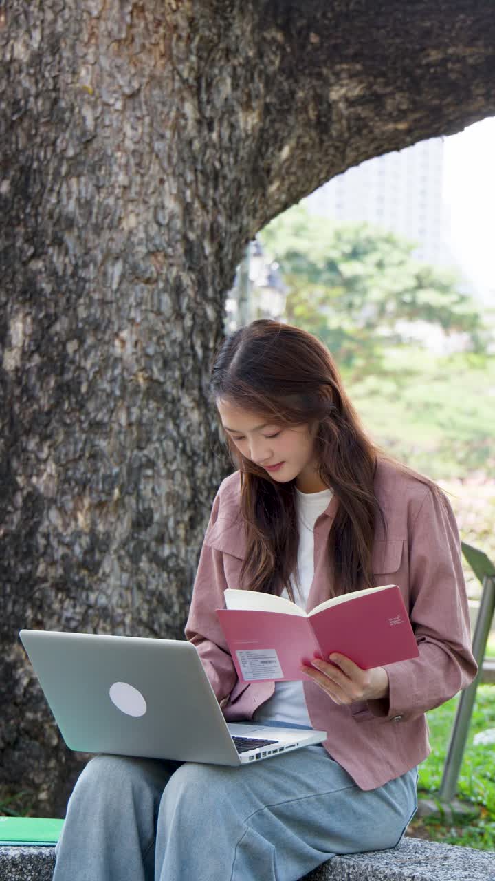 En asiatisk student leser en bok og bruker en laptop mens han/hun sitter under et tre.
