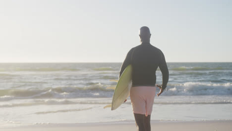 Feliz-Hombre-Afroamericano-Mayor-Caminando-Y-Sosteniendo-Tabla-De-Surf-En-La-Playa,-En-Cámara-Lenta