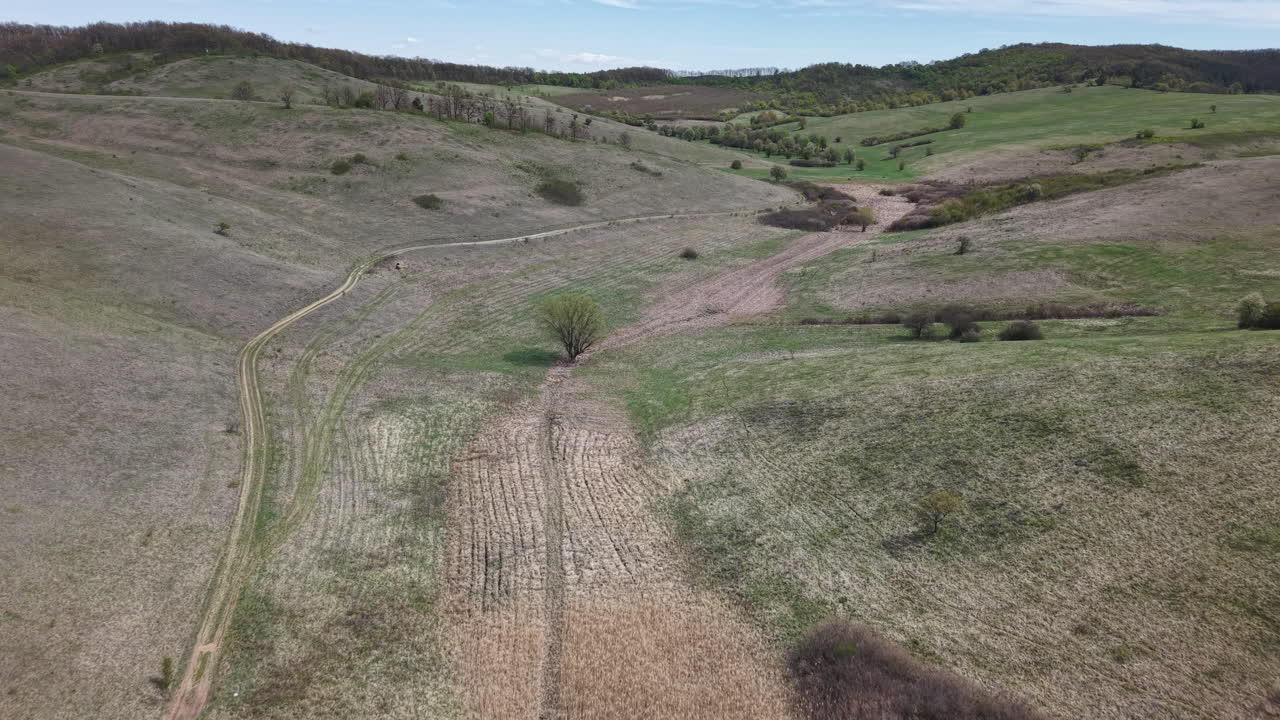 Letecký pohled zachycuje rozsáhlou venkovskou krajinu s zvlněnými kopci, klikatými polními cestami a suchou vegetací pod jasně modrou oblohou, evokující pocit klidné samoty v přírodě.