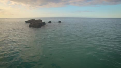 Drone-Volando-Al-Atardecer-Sobre-El-Mar-En-Calma-Hacia-Algunas-Rocas-Desde-Las-Que-Vuela-Un-Pelícano