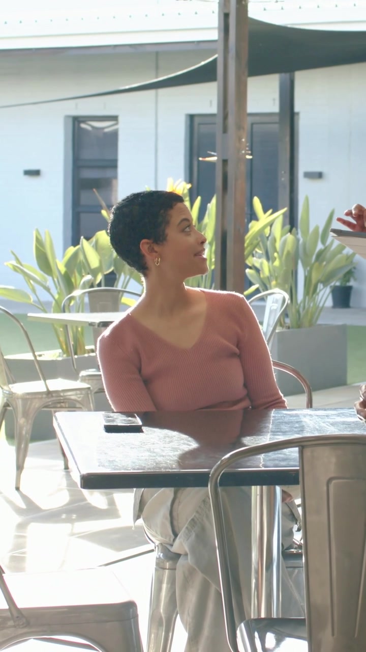 Video verticale: Donna con un top rosa che si gira, capendo l'approccio giusto nel patio di un caffè, prendendo un piatto e il telefono.
