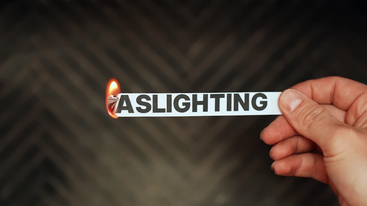 Hand holds a paper strip reading “GASLIGHTING” as the edge burns in flame, symbolizing psychological manipulation, toxic relationships, and emotional abuse. Concept b-roll