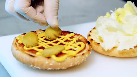 Detailed preparation of delicious hamburger. Chef&#039;s hand putting pickled cucumber parts on a burger bun. Making cheeseburger in fast food restaurant. Close-up