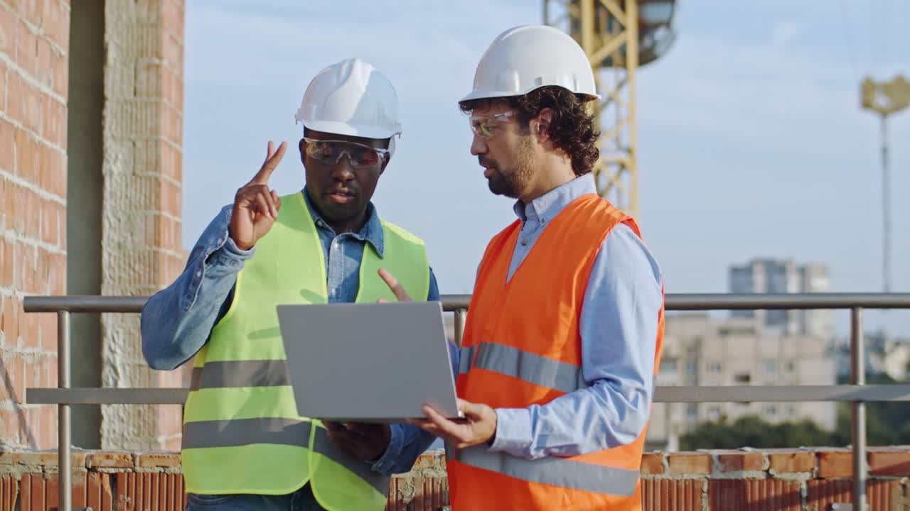 Africa American young male builder and Caucasian architect in hardhats standing at the roof of the building site with a laptop computer and talking about their work.