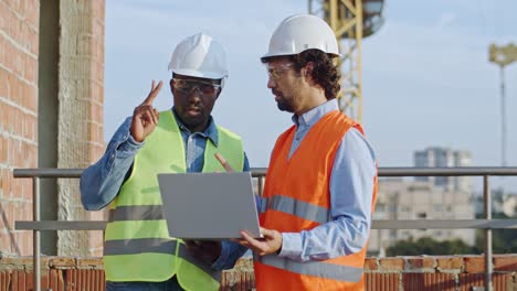 Un-Joven-Constructor-Afroamericano-Y-Un-Arquitecto-Caucásico-Con-Cascos-Parados-En-El-Techo-Del-Sitio-De-Construcción-Con-Una-Computadora-Portátil-Y-Hablando-De-Su-Trabajo.