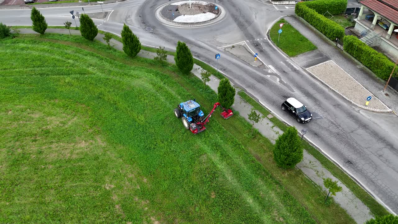 High angle view revealing farmer mowing grass with tractor near roundabout in residential area of Piacenza, showcasing agricultural work amid urban Italian landscape
