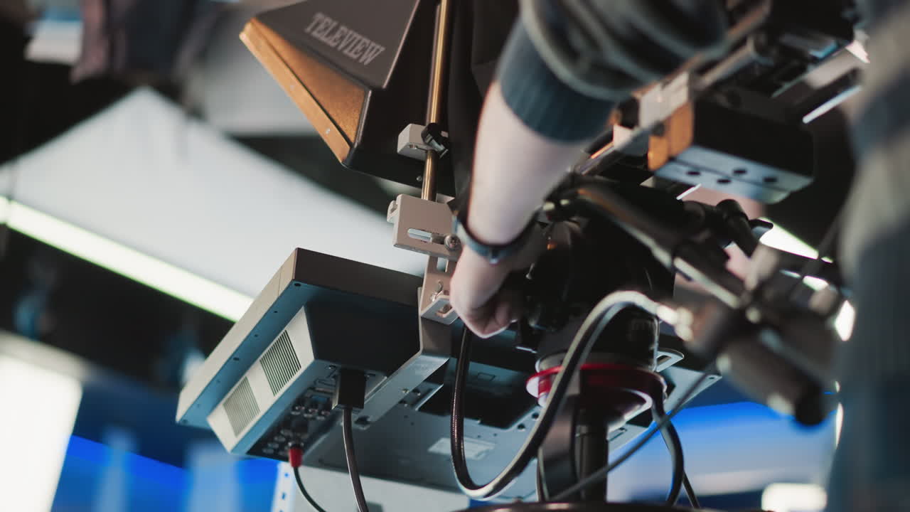 Close-up of hand adjusting professional camera rig with teleprompter in broadcast studio. Operator making fine adjustments to camera while controlling various settings in studio setup
