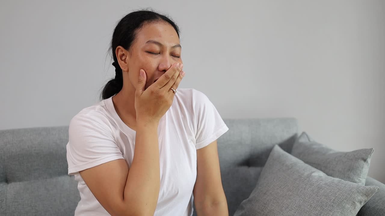 リビングルームのソファに座りながら、口を覆ってあくびをし、腕を伸ばして伸びをする、疲れて眠そうなアジア人女性。