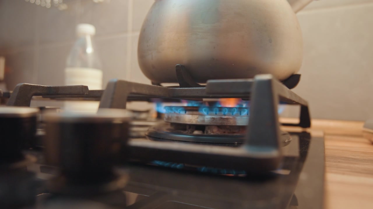 Bollitore in metallo posizionato sul fornello, fiamma blu visibile sotto il bollitore in acciaio inossidabile, acqua che si scalda per tè o ricette, piano di lavoro in legno e paraschizzi piastrellato, proprietario di casa che prepara la bevanda mattutina, atmosfera calda cinematografica