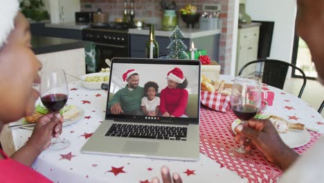 African-american-couple-with-wine-using-laptop-for-christmas-video-call-with-happy-family-on-screen