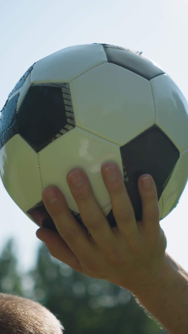 Close-up of a middle-aged man holding a soccer ball high above his head against a clear blue sky