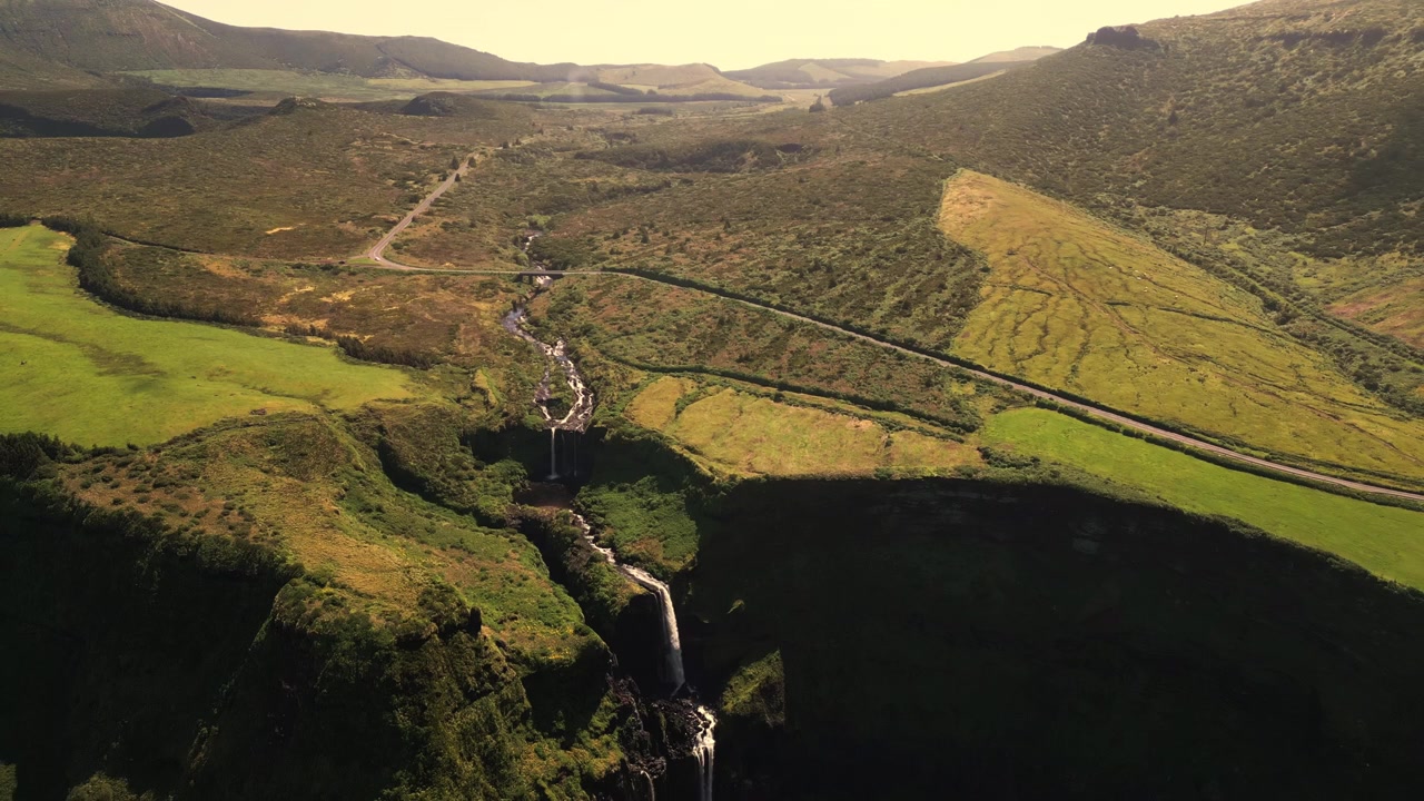 Vista aérea de uma cachoeira em cascata fluindo por colinas verdes e estradas rurais em Flores.