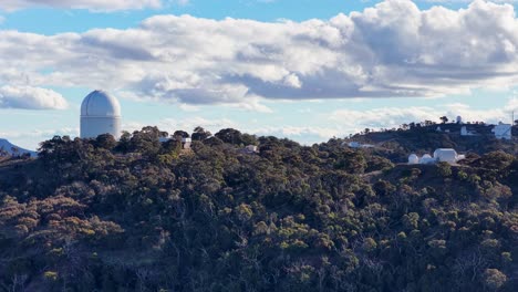 aerial drone footage gradually rises above tree-covered hills, revealing a large white observatory dome under partly cloudy daylight skies in a remote natural landscape