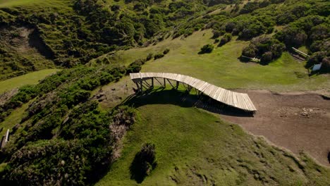 Muelle-de-Las-Almas-on-a-green-hillside-in-Cucao,-Chile-under-clear-blue-skies,-aerial-view