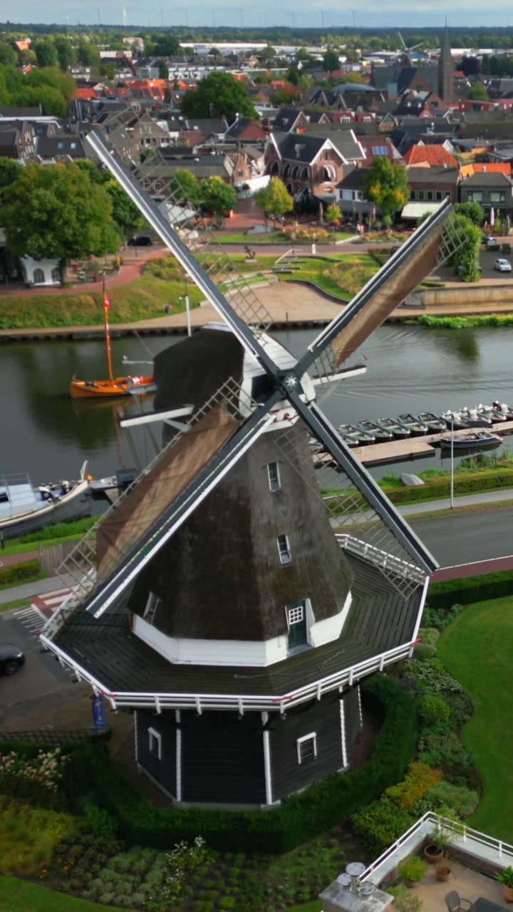 High angle view captures a classic Dutch windmill standing prominently beside a tranquil canal with boats. The charming town spreads behind, showcasing characteristic architecture and green spaces ...