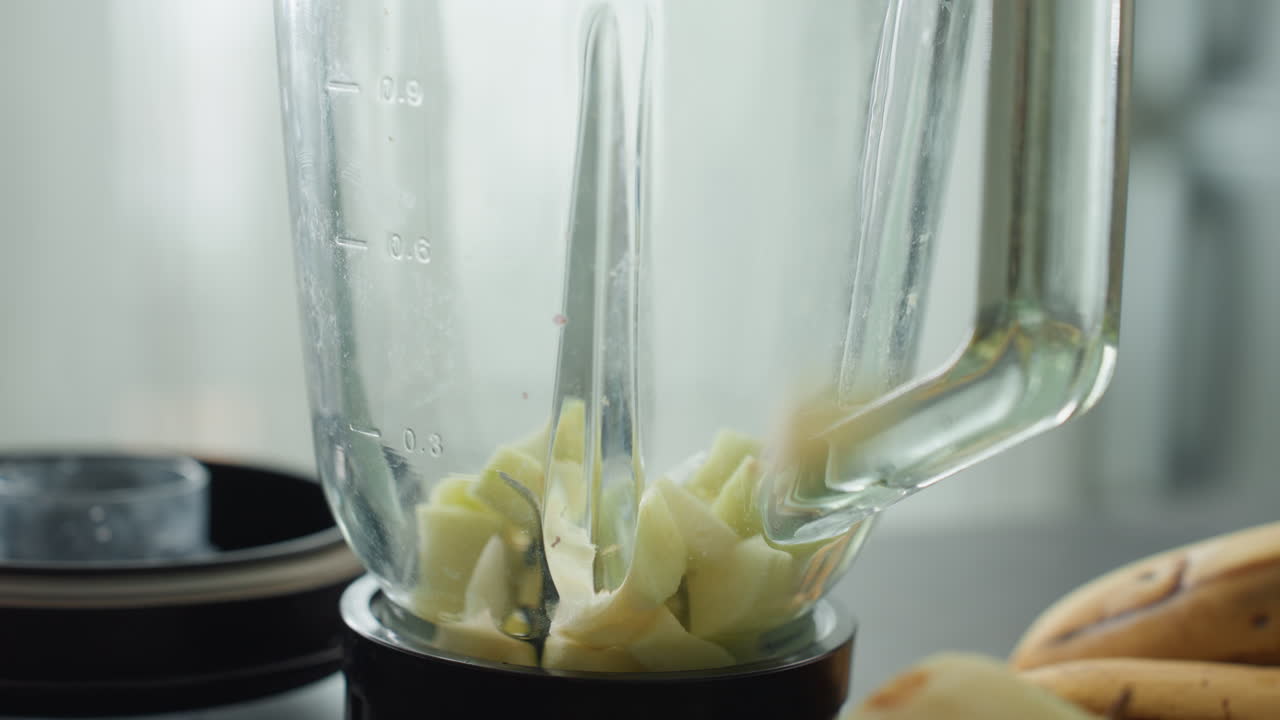 Gravid person blender eplebiter i blenderkanne, nærbilde av hakket frukt som legger seg i bunnen med banan i nærheten, motor klar for pulsfunksjon, morgenlys på kjøkkenet, laging av sunn drikke for gravide.