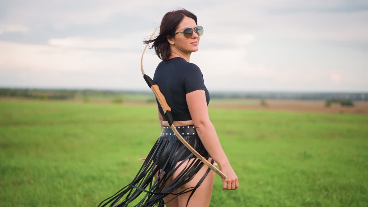 Una donna caucasica, arciere, che cammina in un campo con arco, tracolla e gonna con frange in pelle, occhiali da sole e postura stabile, crea un'atmosfera da cacciatrice forte, ampio prato e orizzonte basso, cielo nuvoloso e delicato, sicura di sé
