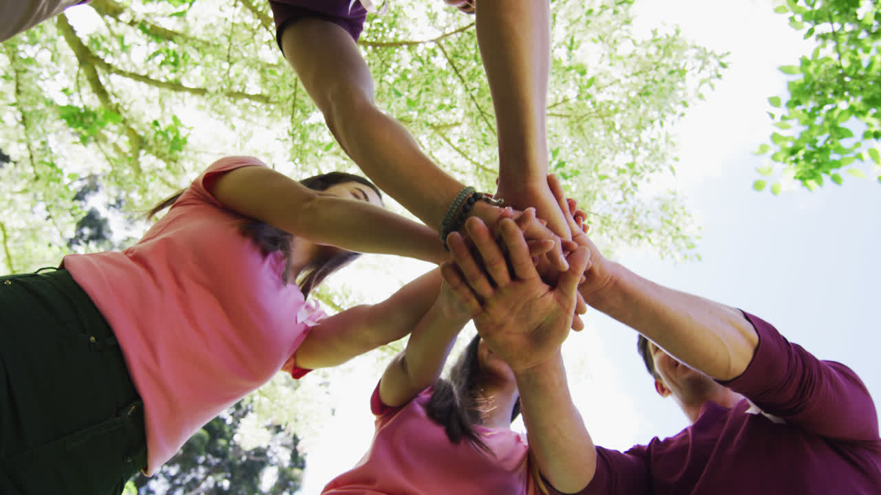 Diverse group of men and women wearing breast cancer ribbons, stacking hands and laughing in park