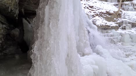 Reducir-A-La-Mitad-La-Cascada-Congelada-Que-Fluye-De-Una-Montaña-En-Invierno,-Suelo-Helado---Toma-De-Revelación-De-Deslizamiento-Lento