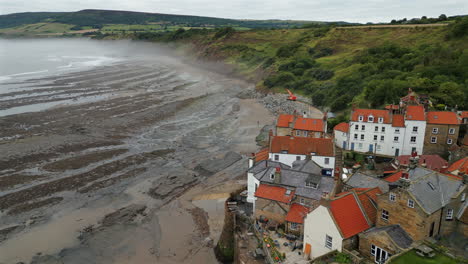 rising establishing aerial drone shot over robin hoods bay looking out to sea on misty morning at low tide