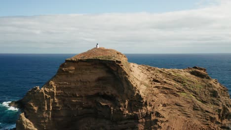 Small-lighthouse-Farol-do-Porto-Moniz-stands-alone-on-island-Ilhéu-Mole,-aerial