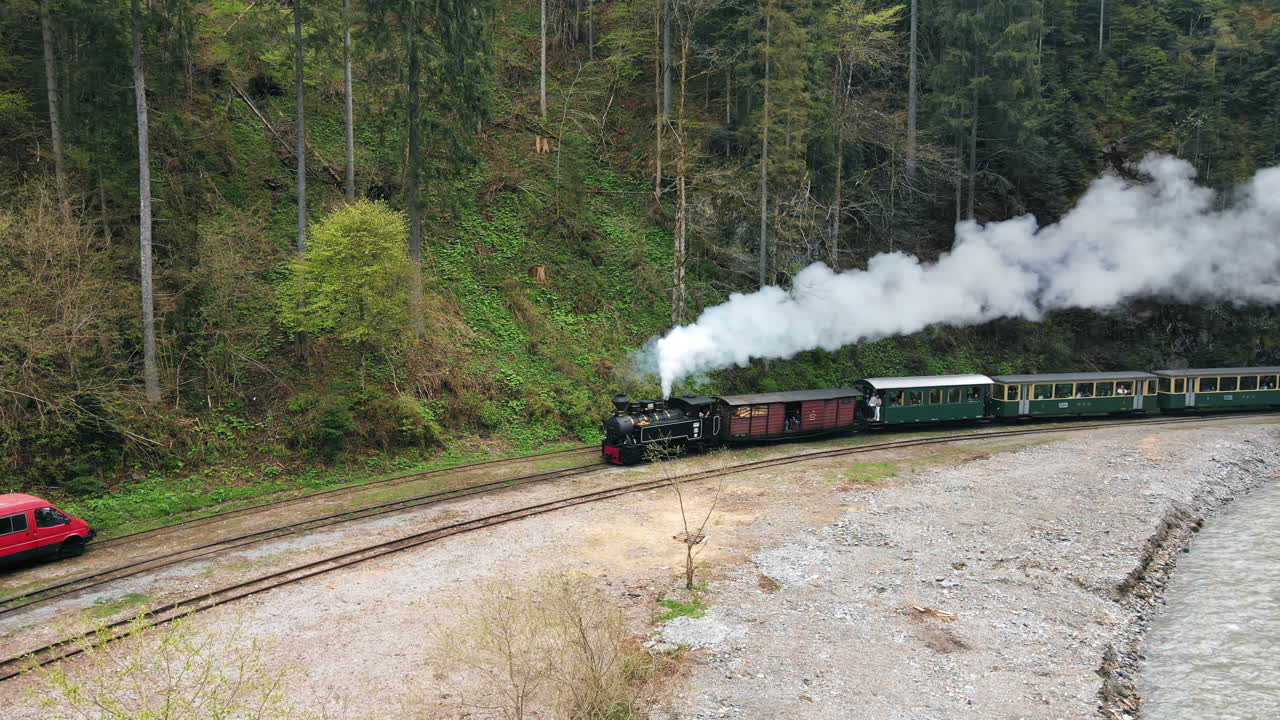 Vista aerea da drone di un treno a vapore in movimento, Mocanita, in una valle con passeggeri a bordo, foresta rigogliosa e fiume intorno, Romania