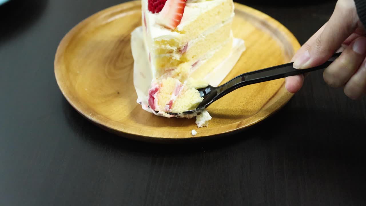 Hand uses black spoon to scoop strawberry shortcake slice on wooden plate, soft indoor lighting