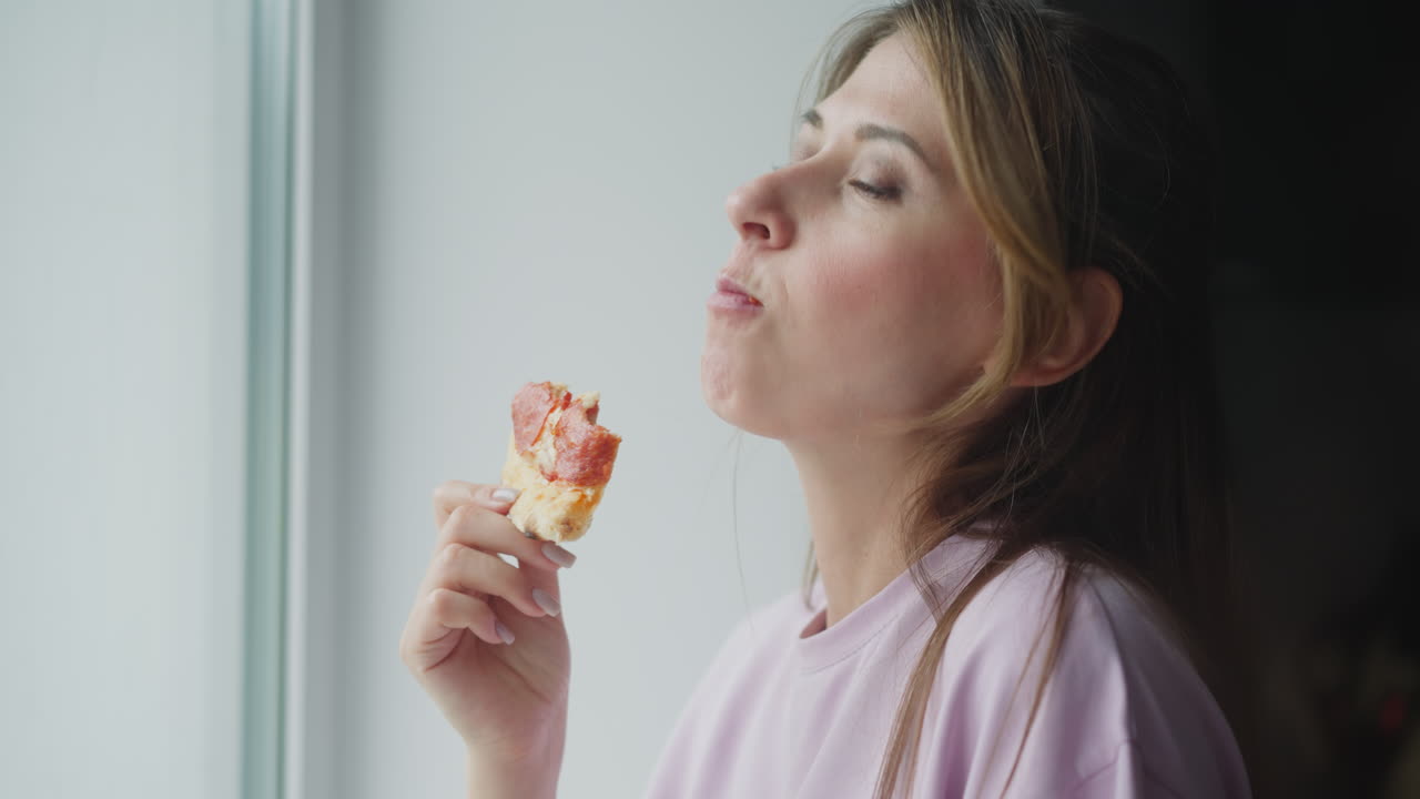 Freelancer eats pizza close to window looking outside through glass, savoring delicious slice while wearing casual light purple shirt, soft natural daylight brightening face with calm cozy mood indoors