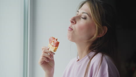 freelancer eats pizza close to window looking outside through glass, savoring delicious slice while wearing casual light purple shirt, soft natural daylight brightening face with calm cozy mood indoors