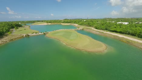 Dynamic-drone-flight-over-tropical-golf-course-with-waving-flag-surrounded-by-lake-during-sunny-day---PLAYA-NUEVA-ROMANA,-Dominican-Republic